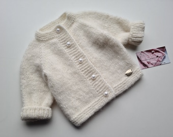 May include: A white knitted baby cardigan with pearl buttons. The cardigan has long sleeves and a button-up front.