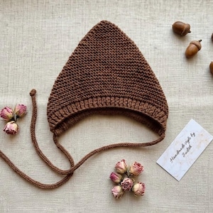 Può includere: Un cappello da folletto marrone lavorato a maglia con lacci, esposto con rose secche e ghiande. Il cappello ha una parte superiore a punta e un bordo a coste. È presente anche una carta con il testo "Handmade style by Judith".