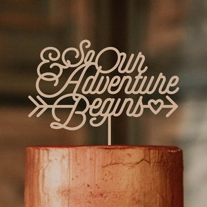 May include: A three-tiered wedding cake with a rustic wooden base. The top tier is a copper-colored cake with a wooden topper that reads "& So Our Adventure Begins". The middle tier is white with a swirled design and the bottom tier is a wooden slice.