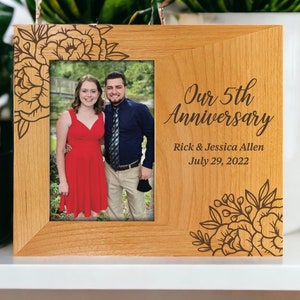 May include: A wooden photo frame with a floral design engraved on the top and bottom. The frame has a photo of a couple in the center. The text "Our 5th Anniversary" is engraved on the frame, along with the names "Rick & Jessica Allen" and the date "July 29, 2022".