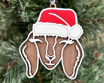 Könnte beinhalten: Weihnachtsbaumschmuck aus Holz in Form einer Ziege, die einen roten und weißen Weihnachtsmann-Hut trägt. Die Ziege hat braunes Fell und weiße Augen.