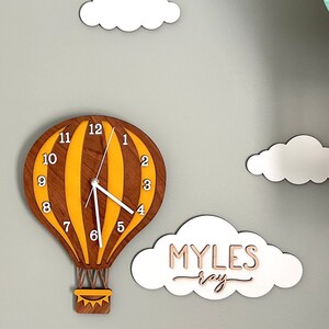 May include: A wooden hot air balloon clock with yellow and brown stripes, white clock hands, and a white cloud with the name "MYLES ray" written on it. There are other white clouds in the background.