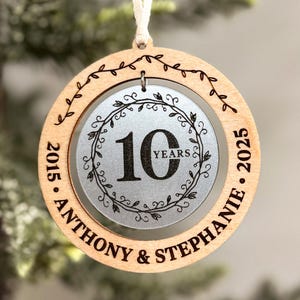 May include: A round wooden ornament with a silver circle in the centre. The silver circle has a wreath design and the number 10 with the word "YEARS" below it. The outer wooden circle has the text "2015 - ANTHONY & STEPHANIE - 2025" around the edge.