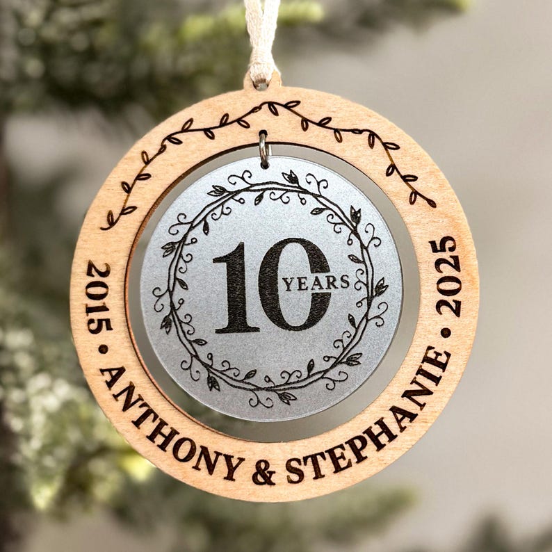 May include: A round wooden ornament with a silver circle in the centre. The silver circle has a wreath design and the number 10 with the word "YEARS" below it. The outer wooden circle has the text "2015 - ANTHONY & STEPHANIE - 2025" around the edge.