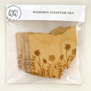 Puede incluir: Conjunto de cuatro posavasos de madera con un dise&ntilde;o grabado con l&aacute;ser del estado de Ohio con flores silvestres. Los posavasos est&aacute;n empaquetados en una bolsa de pl&aacute;stico transparente con una etiqueta blanca que dice "WOODEN COASTER SET".