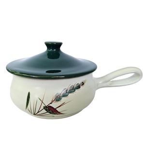 May include: A ceramic soup bowl with a dark green lid and handle. The bowl is off-white with a painted floral design in shades of green, red, and brown. The handle is a loop shape, and the lid has a rounded knob.