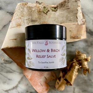 May include: A black-lidded jar of Willow & Birch Relief Salve from Twin Falls Botanicals. The label features text and leaf illustrations. The jar is set on a background of birch bark and other natural elements. The product is 2 oz.