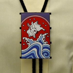 May include: A black bolo tie with a rectangular pendant featuring a red and white stylized image of two rabbits jumping over blue and white waves.