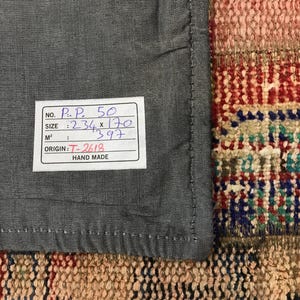 Puede incluir: Primer plano de una tela gris con un borde cosido, junto a una secci&oacute;n de una alfombra colorida y estampada. Una etiqueta blanca con texto negro indica que el tama&ntilde;o es 234 x 170, y el origen es T-2618. La etiqueta tambi&eacute;n dice "Hand Made".
