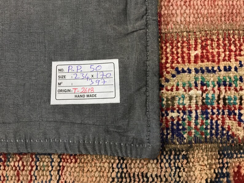 Puede incluir: Primer plano de una tela gris con un borde cosido, junto a una secci&oacute;n de una alfombra colorida y estampada. Una etiqueta blanca con texto negro indica que el tama&ntilde;o es 234 x 170, y el origen es T-2618. La etiqueta tambi&eacute;n dice "Hand Made".