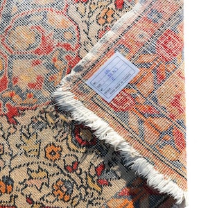 May include: A close-up of a beige, red, and blue patterned rug with a white fringe. The rug has a tag attached that reads "No.: 141, Size: 93 x 63, Origin: F, Hand Made in Turkey."