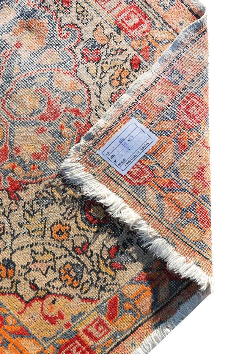 May include: A close-up of a beige, red, and blue patterned rug with a white fringe. The rug has a tag attached that reads "No.: 141, Size: 93 x 63, Origin: F, Hand Made in Turkey."