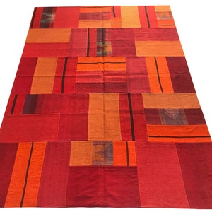 Puede incluir: Una alfombra de patchwork con un diseño geométrico en tonos rojos y naranjas. La alfombra está hecha de tejido tejido y tiene un aspecto rústico y artesanal.