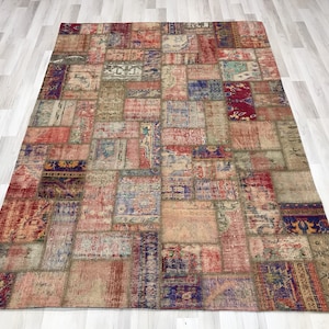 Puede incluir: Una alfombra grande y rectangular de patchwork con un aspecto envejecido. La alfombra presenta una variedad de parches rectangulares y cuadrados en tonos de rojo, azul, beige y morado. La alfombra est&aacute; colocada sobre un suelo de madera clara.