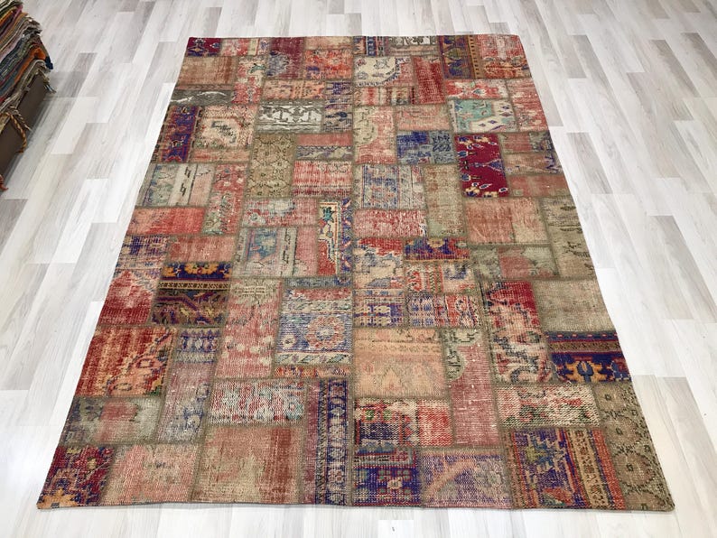 Puede incluir: Una alfombra grande y rectangular de patchwork con un aspecto envejecido. La alfombra presenta una variedad de parches rectangulares y cuadrados en tonos de rojo, azul, beige y morado. La alfombra est&aacute; colocada sobre un suelo de madera clara.