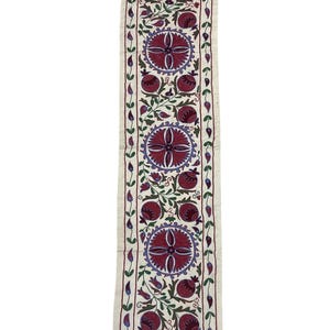Puede incluir: Un camino de mesa de algodón blanco con un patrón repetido de granadas y diseños florales bordados en rojo y morado.