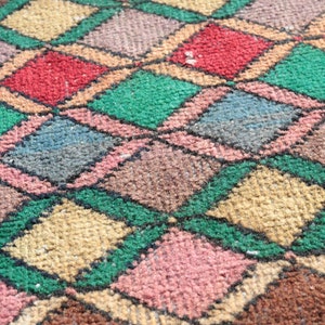 May include: A close-up of a colorful diamond patterned rug with shades of green, brown, pink, blue, and beige. The rug is woven with a textured, fuzzy surface.