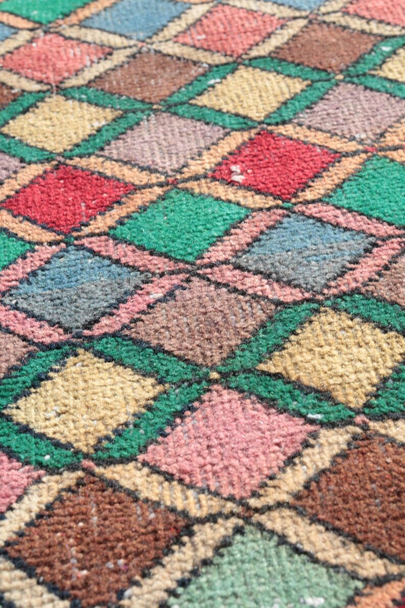 May include: A close-up of a colorful diamond patterned rug with shades of green, brown, pink, blue, and beige. The rug is woven with a textured, fuzzy surface.