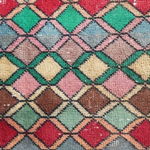 May include: A close-up of a colorful woven rug with a diamond pattern. The rug is made of different shades of pink, red, green, brown, and beige.
