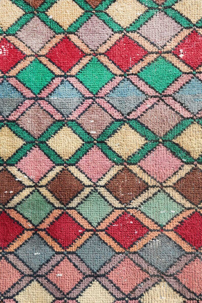 May include: A close-up of a colorful woven rug with a diamond pattern. The rug is made of different shades of pink, red, green, brown, and beige.
