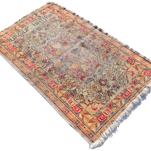 May include: A vintage rug with a floral pattern in shades of red, orange, blue, and beige. The rug has a fringe border and the text "URGUF HATIRA" is woven into the design.