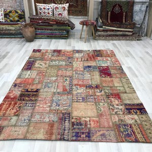 Puede incluir: Una alfombra grande y rectangular de patchwork con un aspecto envejecido y vintage. La alfombra presenta una variedad de parches rectangulares y cuadrados en tonos rojos, azules, beige y morados. La alfombra se exhibe sobre un suelo de madera clara.