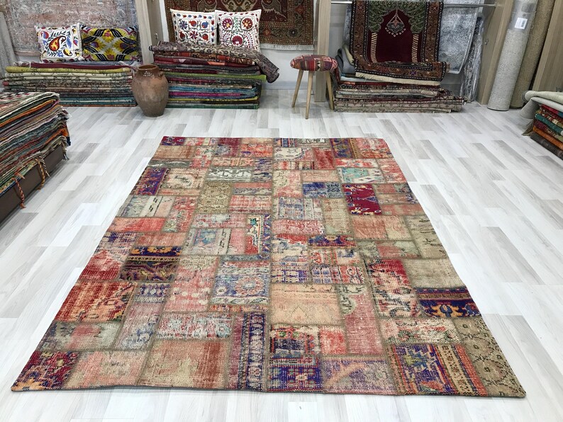 Puede incluir: Una alfombra grande y rectangular de patchwork con un aspecto envejecido y vintage. La alfombra presenta una variedad de parches rectangulares y cuadrados en tonos rojos, azules, beige y morados. La alfombra se exhibe sobre un suelo de madera clara.