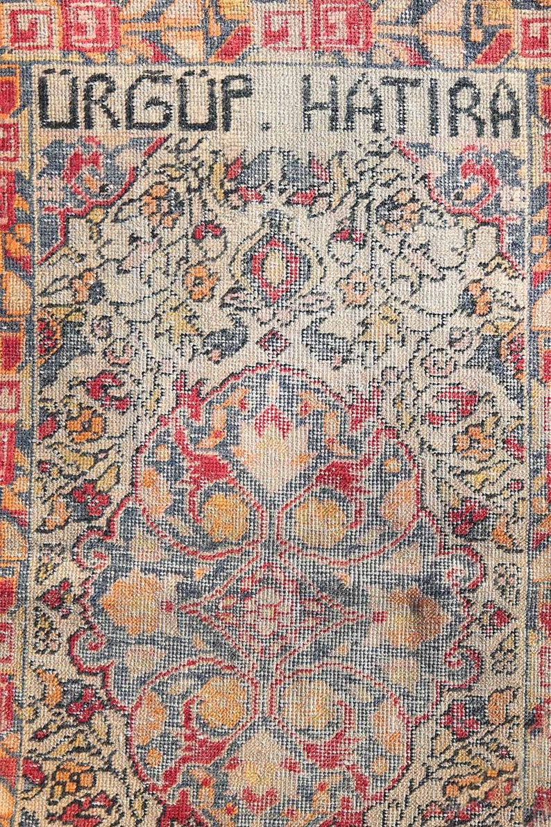 May include: A vintage rug with a faded floral pattern in shades of red, orange, and blue. The rug has a border with a geometric design. The text "URGUP HATIRA" is woven into the rug.