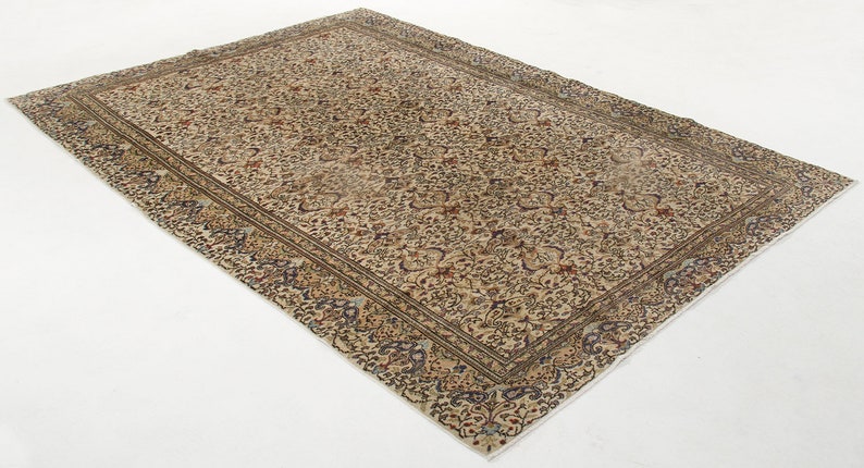 K&ouml;nnte beinhalten: Ein Vintage-Teppich mit cremefarbenem Hintergrund und einem floralen Muster in Braun-, Blau- und Rott&ouml;nen. Der Teppich hat einen Rand mit einem sich wiederholenden Muster.