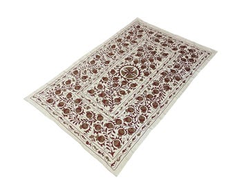 May include: Rectangular off-white textile with a repeating floral pattern in shades of brown and burgundy. The design features stylized flowers and foliage, creating a symmetrical and ornate composition. The textile has a border with a similar floral motif.