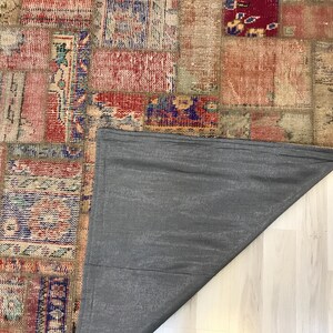 Puede incluir: Una alfombra patchwork con un aspecto envejecido y vintage. La alfombra presenta una variedad de parches rectangulares y cuadrados en tonos de rojo, beige, azul y naranja. La parte trasera de la alfombra es de tela gris lisa.