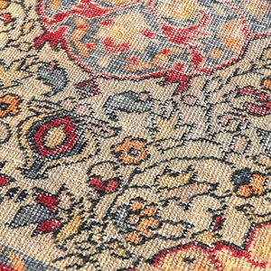 May include: Close-up of a beige rug with a detailed floral pattern in shades of red, orange, yellow, and black. The rug has a woven texture and a fringe border.