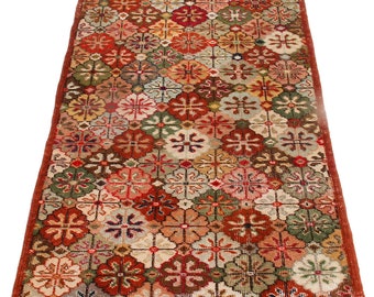 May include: A rectangular rug with a repeating floral pattern in shades of red, green, orange, and cream. The rug has a dark red border and a fringed edge. The design features a geometric arrangement of stylized flowers.