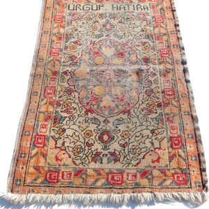 May include: A vintage Turkish rug with a floral pattern in shades of red, orange, blue, and beige. The rug has a fringe border and the text "URGUF HATIRA" is woven into the top border.