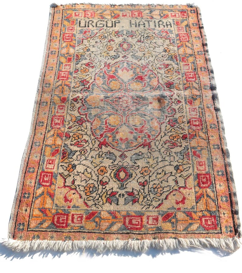 May include: A vintage Turkish rug with a floral pattern in shades of red, orange, blue, and beige. The rug has a fringe border and the text "URGUF HATIRA" is woven into the top border.