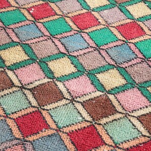 May include: A close-up of a colorful woven rug with a geometric pattern of squares and diamonds in shades of red, green, brown, beige, pink, and blue.