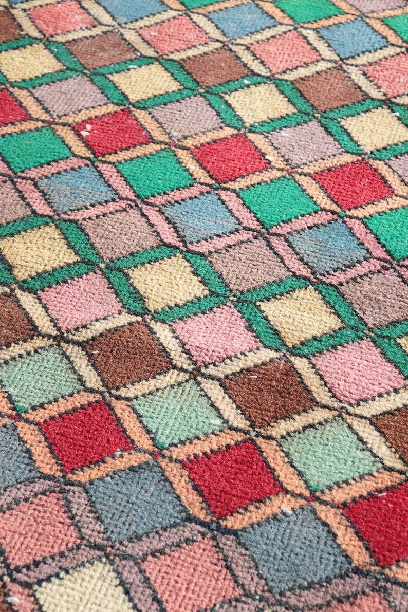 May include: A close-up of a colorful woven rug with a geometric pattern of squares and diamonds in shades of red, green, brown, beige, pink, and blue.
