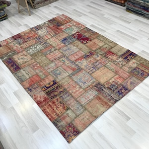 Puede incluir: Una alfombra rectangular de patchwork con un aspecto envejecido y vintage. La alfombra presenta una variedad de parches rectangulares en tonos rojos, azules, beige y morados. La alfombra está colocada sobre un suelo de madera clara.