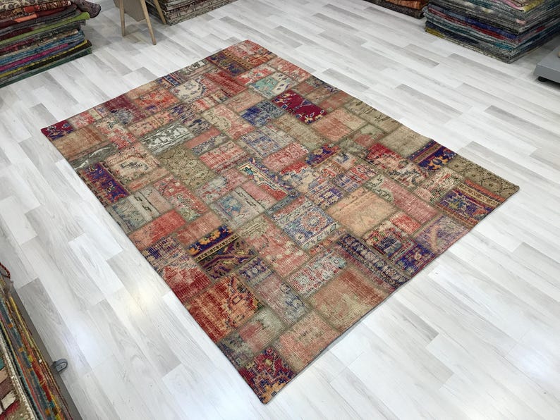 Puede incluir: Una alfombra rectangular de patchwork con un aspecto envejecido y vintage. La alfombra presenta una variedad de parches rectangulares en tonos rojos, azules, beige y morados. La alfombra est&aacute; colocada sobre un suelo de madera clara.