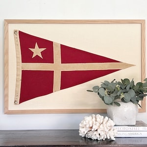 May include: A framed vintage nautical flag with a red background, a white star, and a beige cross. The flag is displayed on a wall above a shelf with books and a white vase with green foliage.