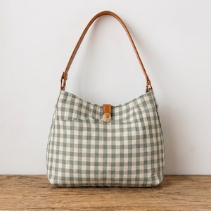 May include: A hobo-style shoulder bag featuring a sage green and white gingham check pattern. The bag includes a single brown leather strap attached with metal hardware and a central brown leather tab closure secured with a brass-toned metal snap button.