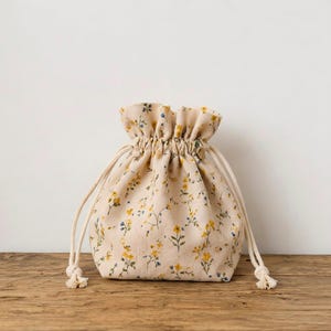 May include: A small drawstring pouch made of off-white fabric featuring a repeating pattern of tiny yellow flowers and small blue leaves. The bag has a gathered top opening secured by two thick, cream-coloured braided cotton cords on each side.