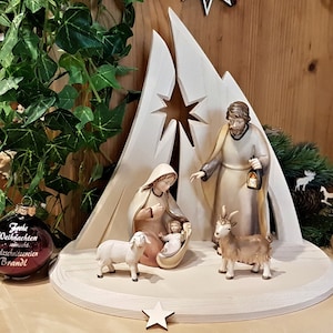 Könnte beinhalten: Eine hölzerne Weihnachtskrippe mit einem Stern-Ausschnitt, die Figuren von Maria, Josef und dem Jesuskind zeigt. Die Szene enthält auch ein Lamm und eine Ziege. Die Figuren sind in hellem Braun bemalt und die Szene steht auf einem weißen Holzsockel.