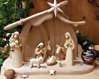 May include: A wooden nativity scene with figurines of Mary, Joseph, baby Jesus, a donkey, a cow, and two sheep. The scene is set in a wooden stable with a star on top. There is a red Christmas ornament with the text "Frohe Weihnachten" and "Heidelattern Brandl" on it.