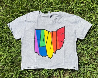 Può includere: Un crop top grigio con un contorno arcobaleno dell'Ohio e la parola "LOVE" al centro. La maglietta è realizzata in un materiale morbido ed è appoggiata sull'erba verde.