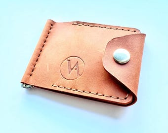 May include: A light brown leather money clip wallet with a snap closure. The wallet has a stitched border and a silver-colored money clip. A circular emblem with a stylized letter "W" is embossed on the front.