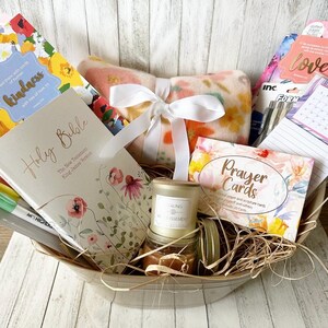 May include: A gift basket filled with items for relaxation and self-care. The basket includes a blanket, a candle, a book of prayer cards, a journal, a Holy Bible, and pens. The basket is lined with straw and has a white ribbon tied around it.