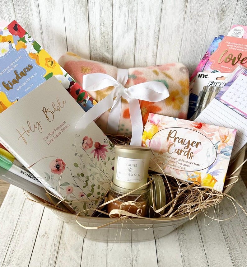 May include: A gift basket filled with items for relaxation and self-care. The basket includes a blanket, a candle, a book of prayer cards, a journal, a Holy Bible, and pens. The basket is lined with straw and has a white ribbon tied around it.