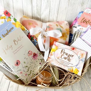 May include: A gift basket filled with items for relaxation and inspiration. It includes a floral patterned throw blanket, a Holy Bible, a book of prayer cards, a candle, and a set of pens.