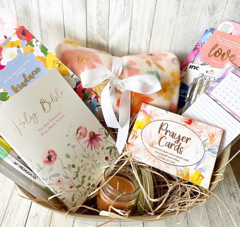 May include: A gift basket filled with items for relaxation and inspiration. It includes a floral patterned throw blanket, a Holy Bible, a book of prayer cards, a candle, and a set of pens.